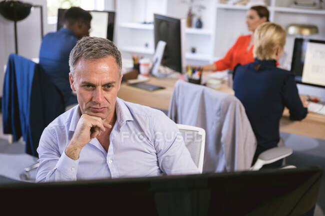 Um homem de negócios caucasiano trabalhando em um escritório moderno, sentado em uma mesa e usando um computador, com seus colegas de negócios trabalhando em segundo plano — Fotografia de Stock