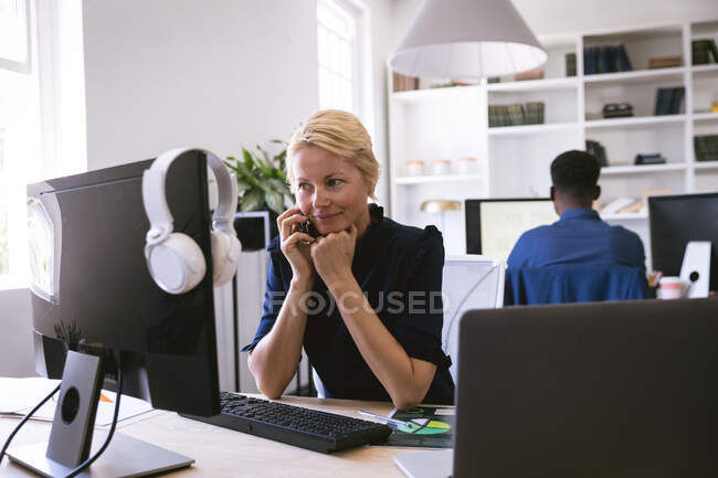 Una mujer de negocios caucásica trabajando en una oficina moderna, sentada en un escritorio y usando una computadora, hablando en un teléfono inteligente, con sus colegas de negocios trabajando en segundo plano - foto de stock