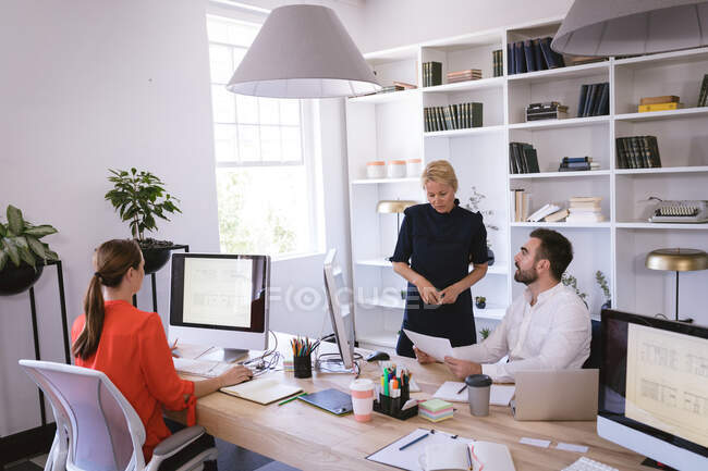 Grupo multiétnico de colegas de negocios masculinos y femeninos que trabajan en una oficina moderna, sentados en un escritorio, usando computadoras, discutiendo su trabajo - foto de stock