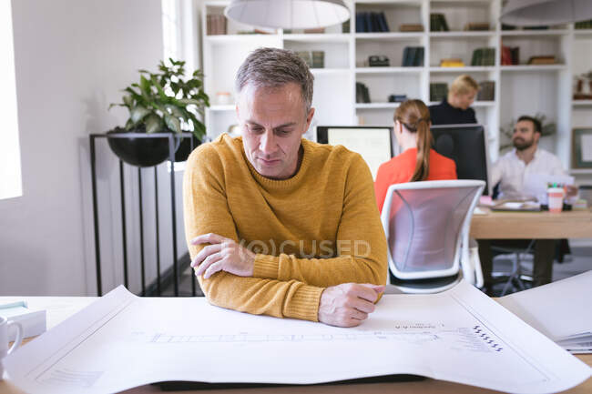 Un uomo d'affari caucasico che lavora in un ufficio moderno, seduto a una scrivania e guarda i piani, con i suoi colleghi di lavoro che lavorano sullo sfondo — Foto stock