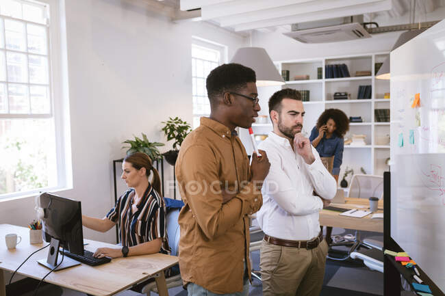Un afroamericano y un hombre de negocios caucásico trabajando en una oficina moderna, mirando una pizarra blanca y una lluvia de ideas juntos, con sus colegas de negocios trabajando en segundo plano - foto de stock