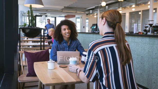 Una raza mixta y una mujer de negocios caucásica trabajando en una oficina moderna, sentada en el comedor de su oficina, bebiendo café y hablando, una mujer usando una computadora portátil - foto de stock