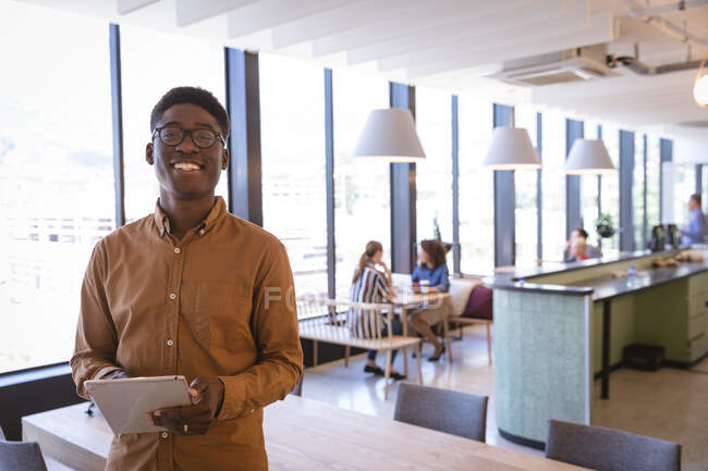 Porträt eines afroamerikanischen Geschäftsmannes, der in einem modernen Büro arbeitet, stehend, ein Tablet in der Hand, in die Kamera blickend und lächelnd, während seine Geschäftskollegen im Hintergrund sitzen. — Stockfoto