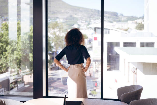 Вид спереду змішаної гонки бізнес-леді, що працює в сучасному офісі, стоїть і дивиться з вікна мислення, з руками на стегнах, в сонячний день — стокове фото