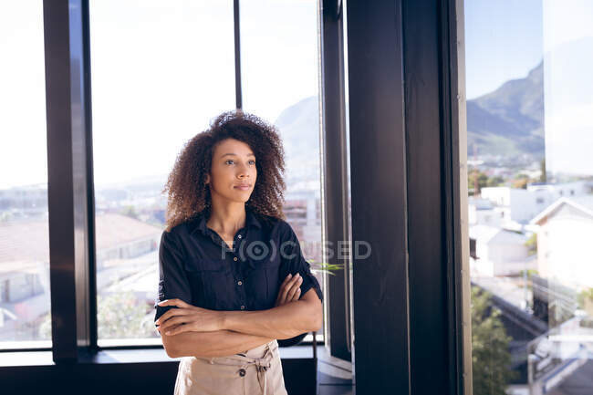 Деловая женщина смешанной расы, работающая в современном офисе, стоящая у окна, скрещивающая руки и думающая, в солнечный день — стоковое фото