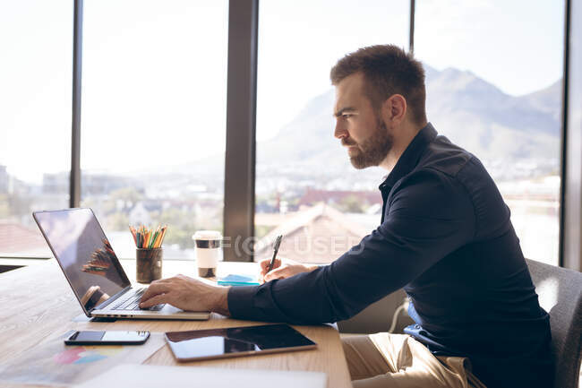 Вид сбоку на кавказского бизнесмена, работающего в современном офисе, сидящего за столом у окна, пользующегося ноутбуком и пишущего ручкой, делающего заметки — стоковое фото