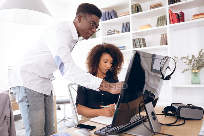 Деловая женщина смешанной расы и афро-американский бизнесмен, работающий в современном офисе, за компьютером и беседующий, мужчина, стоящий и указывающий на экран — стоковое фото