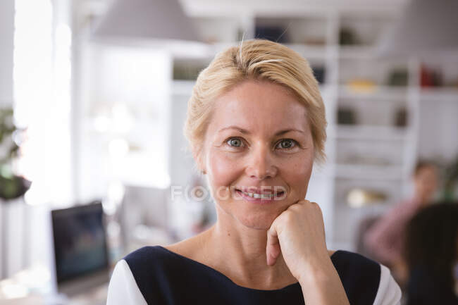 Retrato de una mujer de negocios caucásica feliz trabajando en una oficina moderna, mirando a la cámara y sonriendo, con sus colegas trabajando en el fondo - foto de stock