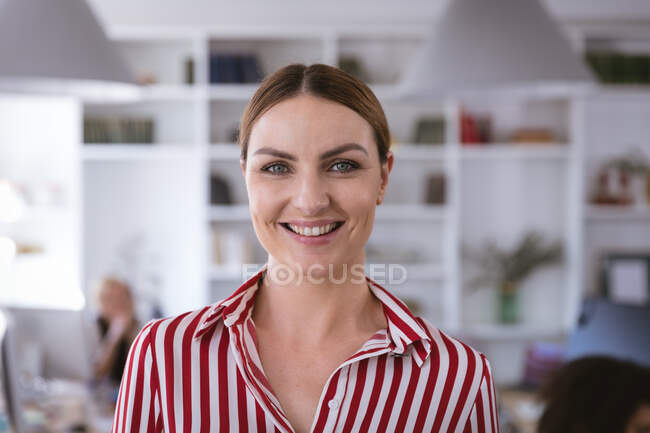 Портрет счастливой кавказской предпринимательницы, работающей в современном офисе, смотрящей в камеру и улыбающейся, с коллегами, работающими на заднем плане — стоковое фото