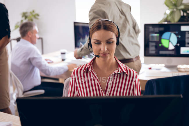 A Caucasian businesswoman working in a modern office, sitting at a desk, using a laptop computer, wearing headset and talking, with her business colleagues working in the background — Stock Photo