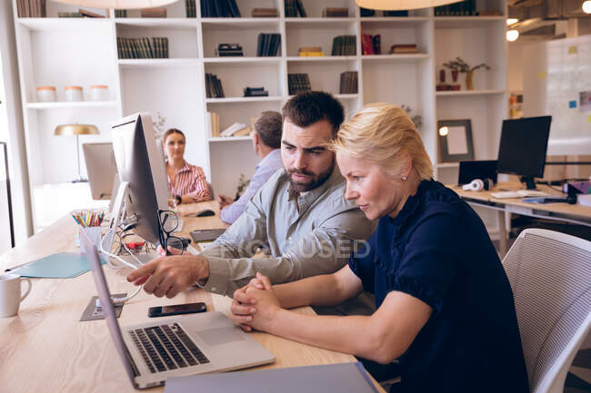 Eine kaukasische Geschäftsfrau und ein Geschäftsmann, die in einem modernen Büro arbeiten, einen Laptop benutzen und miteinander reden, während ihre Geschäftskollegen im Hintergrund arbeiten — Stockfoto