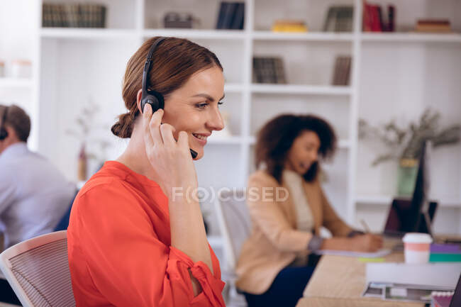 A Caucasian businesswoman working in a modern office, sitting at a desk, using a laptop computer, wearing headset and talking on the phone, with her business colleagues working in the background — Stock Photo
