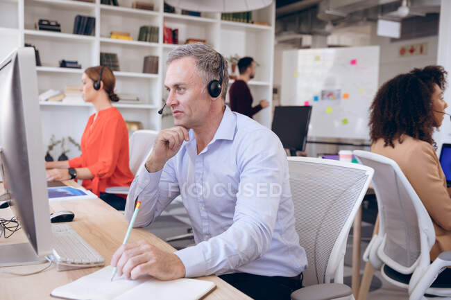 Кавказский бизнесмен, работающий в современном офисе, сидящий за столом, в наушниках и разговаривающий по телефону, со своими коллегами по бизнесу, работающими на заднем плане — стоковое фото