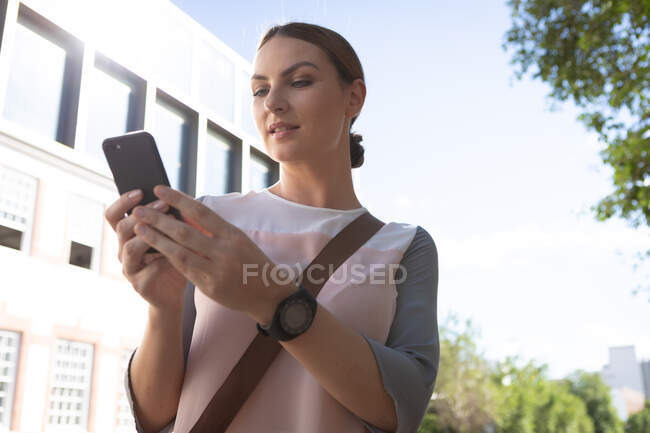 A Caucasian businesswoman on the go on a sunny day, holding and using her smartphone, wearing fashionable clothes and a smartwatch — Stock Photo
