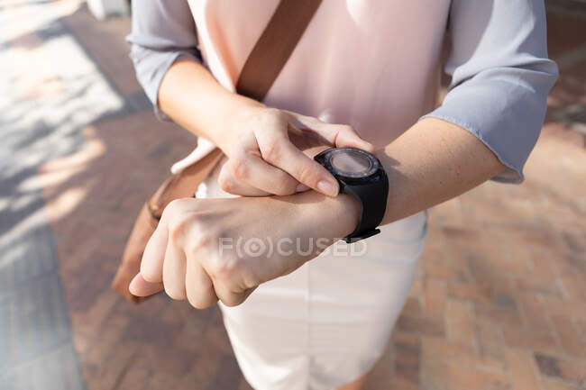 Primer plano de la mujer de negocios sobre la marcha en un día soleado, de pie y comprobando su reloj inteligente, usando ropa de moda - foto de stock