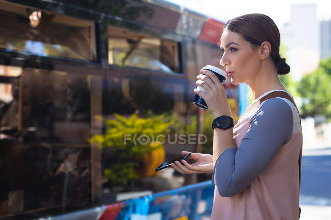 Eine kaukasische Geschäftsfrau an einem sonnigen Tag unterwegs, hält ihr Smartphone vor sich und trinkt einen Kaffee zum Mitnehmen — Stockfoto
