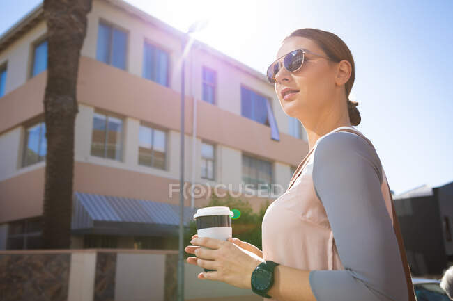 Uma mulher de negócios caucasiana em movimento em um dia ensolarado, de pé e segurando um café takeaway, usando óculos de sol e um smartwatch — Fotografia de Stock