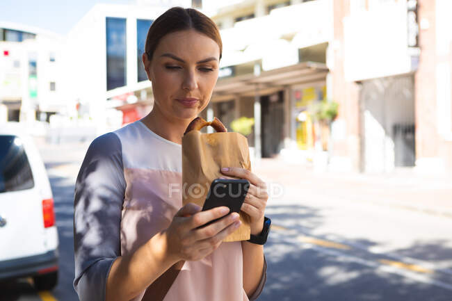 A Caucasian businesswoman on the go on a sunny day, standing on a city street, eating a bagel and texting on her smartphone — Stock Photo