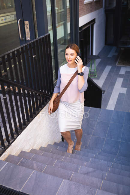 Una mujer de negocios caucásica en movimiento en un día soleado, caminando por las escaleras, hablando en su teléfono inteligente y usando un reloj inteligente - foto de stock