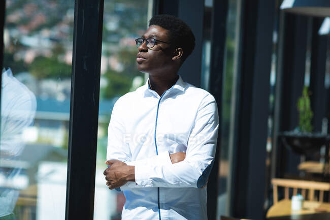 An African American businessman with short hair, wearing a white shirt and glasses standing inside a cafe and looking at the window with arms crossed — Stock Photo