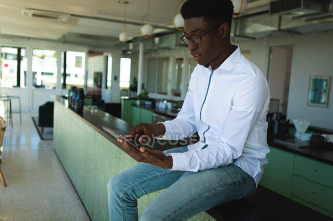 An African American businessman with short hair, wearing a white shirt and glasses sitting on countertop inside a cafe, holding and using his tablet — Stock Photo