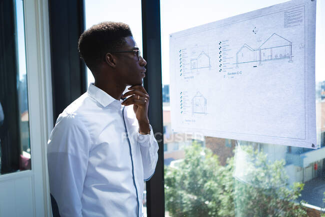 Un homme d'affaires afro-américain, vêtu d'une chemise blanche, travaillant dans un bureau moderne, se tenant par la fenêtre, touchant son menton et regardant les plans sur la fenêtre — Photo de stock