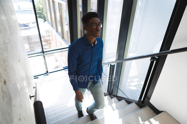 Un hombre de negocios afroamericano de pelo corto oscuro, con camisa y gafas azules, trabajando en una oficina moderna, caminando por las escaleras - foto de stock