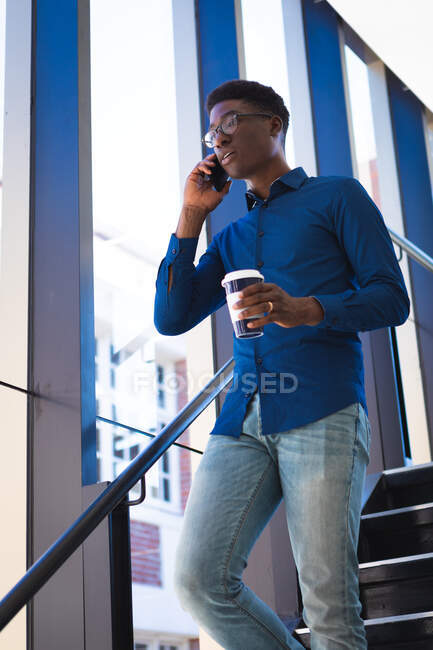Un uomo d'affari afroamericano con i capelli corti scuri, indossa una camicia blu e occhiali, lavora in un ufficio moderno, cammina sulle scale, parla al telefono e tiene in mano un caffè da asporto — Foto stock