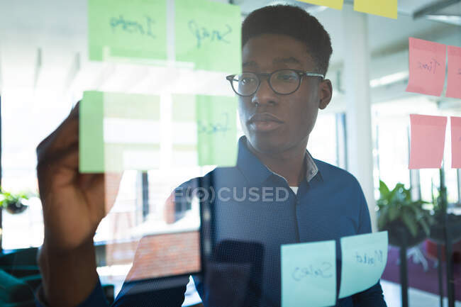 Um empresário afro-americano vestindo uma camisa azul e óculos, trabalhando em um escritório moderno, escrevendo em quadro claro com notas de memorando — Fotografia de Stock