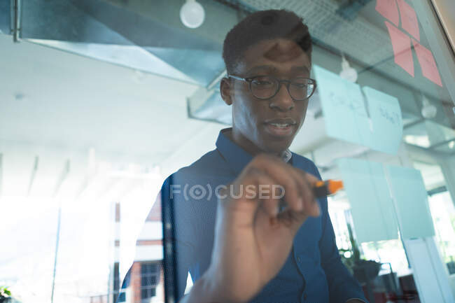 Ein afroamerikanischer Geschäftsmann trägt ein blaues Hemd und eine Brille, arbeitet in einem modernen Büro und schreibt auf Klarsichttafel mit Notizen — Stockfoto