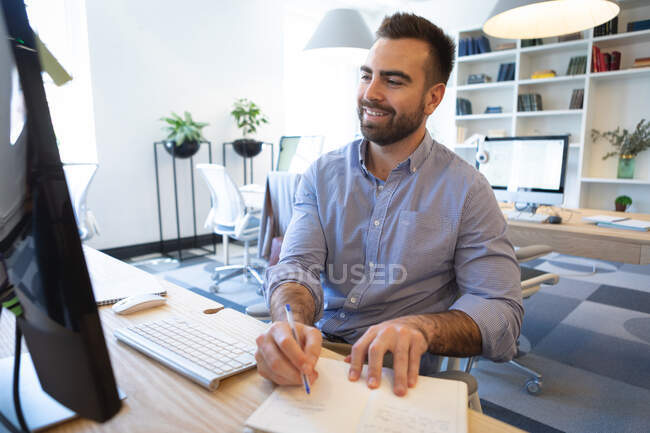 Кавказский бизнесмен с короткими волосами, в синей рубашке, работает в современном офисе, делает заметки и улыбается, используя настольный компьютер — стоковое фото