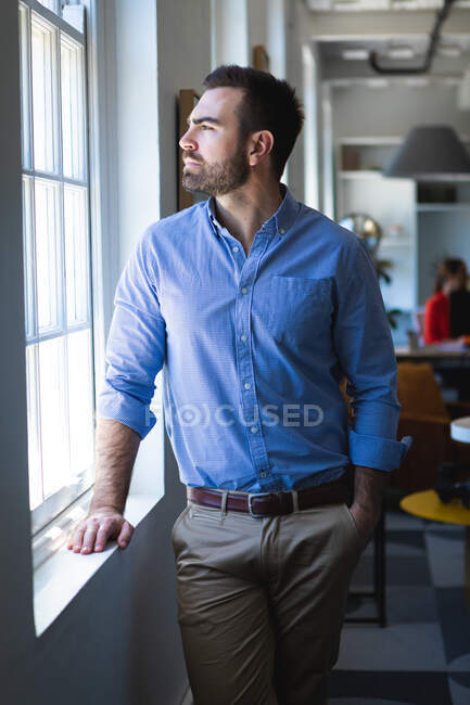 Un hombre de negocios caucásico con el pelo corto, con una camisa azul, trabajando en una oficina moderna, de pie junto a la ventana y mirando a través de la ventana - foto de stock
