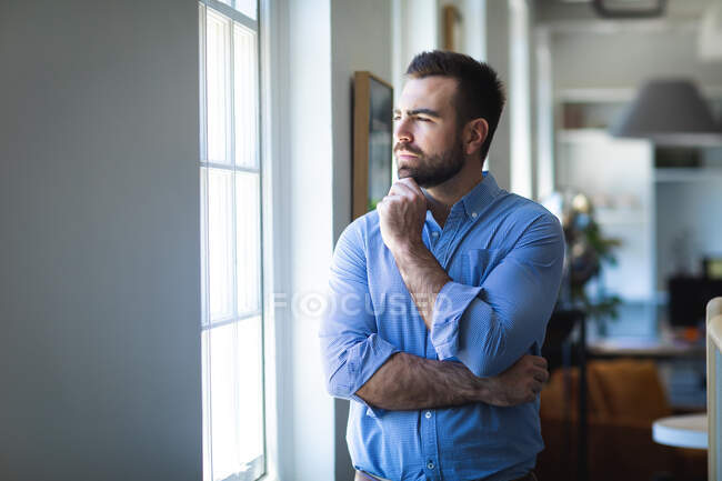 Ein kaukasischer Geschäftsmann mit kurzen Haaren, blauem Hemd, der in einem modernen Büro arbeitet, am Fenster steht und sein Kinn berührt — Stockfoto