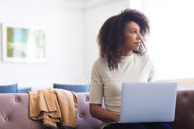 Деловая женщина смешанной расы с вьющимися волосами, работающая в современном офисе, сидящая на диване и работающая на ноутбуке — стоковое фото