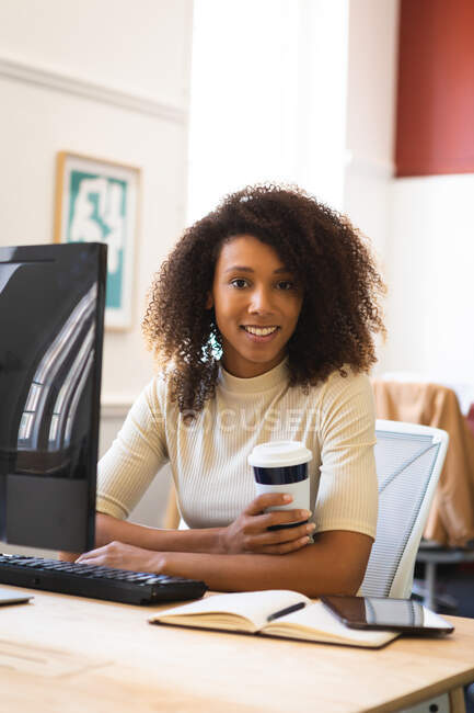 Ritratto di una donna d'affari mista con i capelli ricci, che lavora in un ufficio moderno, siede a un tavolo e tiene in mano un caffè da asporto, guarda la macchina fotografica — Foto stock