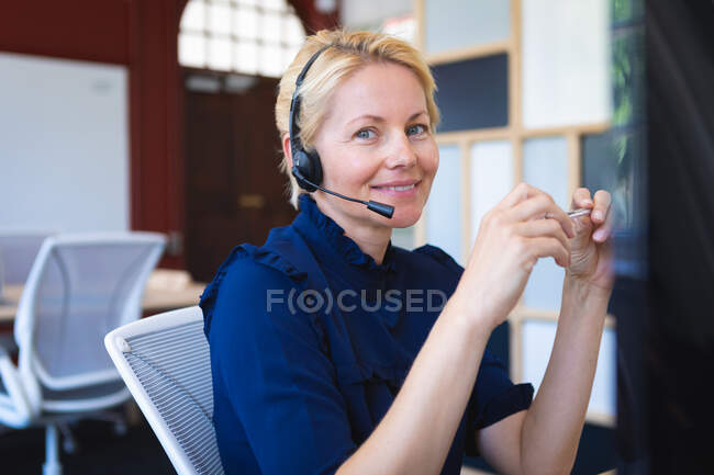 Portrait of a Caucasian businesswoman with short blond hair, working in a modern office, sitting at a desk, wearing headset and looking at camera — Stock Photo