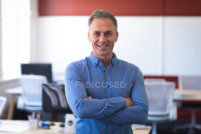 Портрет кавказского бизнесмена в голубой рубашке, стоящего и улыбающегося со скрещенными руками, работающего в современном офисе, смотрящего в камеру — стоковое фото