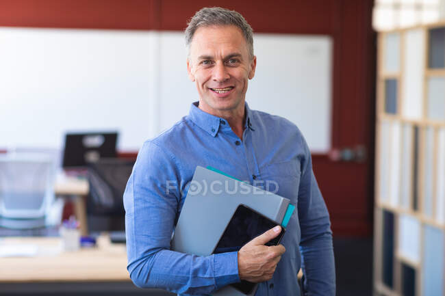 Retrato de un hombre de negocios caucásico con una camisa azul, de pie y sonriente, trabajando en una oficina moderna, mirando a la cámara y sosteniendo su tableta - foto de stock