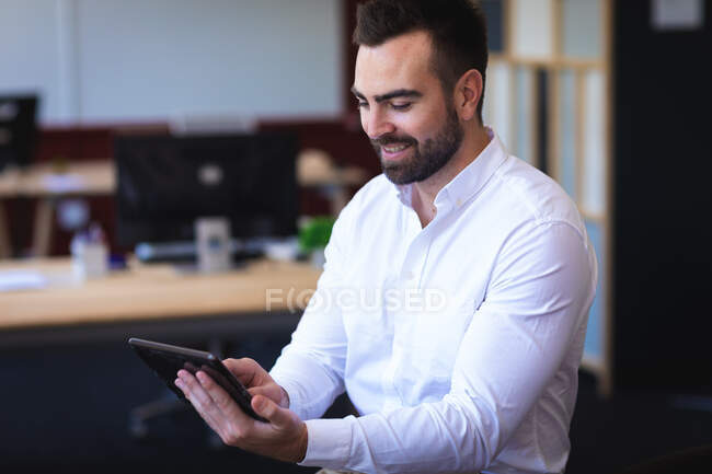 Кавказский бизнесмен в белой рубашке, работает в современном офисе, стоит и улыбается, держит и пользуется планшетом — стоковое фото