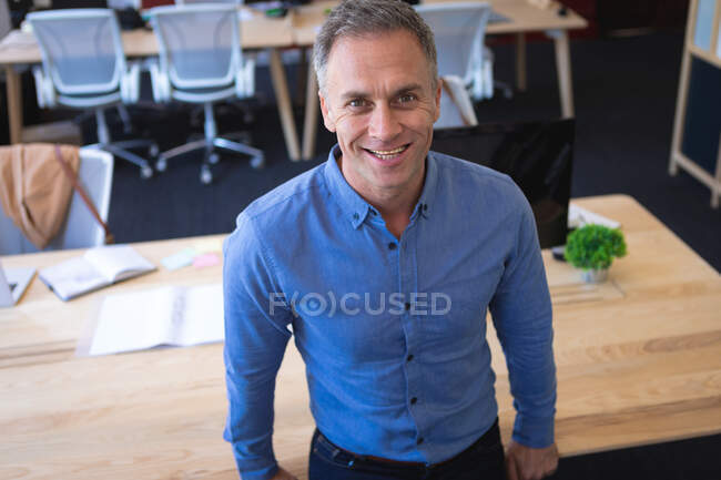 Retrato de un hombre de negocios caucásico con una camisa azul, trabajando en una oficina moderna, de pie y sonriendo, mirando a la cámara - foto de stock