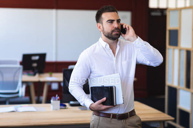 Un hombre de negocios caucásico con una camisa blanca, trabajando en una oficina moderna, de pie y hablando por teléfono, sosteniendo su tableta - foto de stock