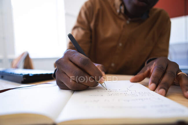 Крупный план афроамериканца, работающего в современном офисе, сидящего за столом и делающего заметки ручкой — стоковое фото