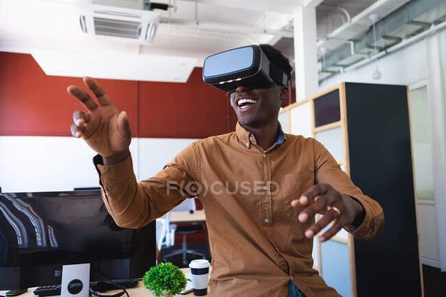 Un hombre afroamericano, trabajando en una oficina moderna, sentado en una mesa y sonriendo, con auriculares VR y tocando la pantalla interactiva virtual - foto de stock
