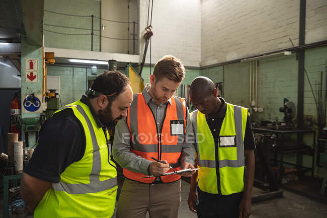 Deux ouvriers blancs et un ouvrier afro-américain portant une veste haute vis parlant et tenant un presse-papiers. Travailleurs de l'industrie dans une usine de fabrication d'équipements hydrauliques. — Photo de stock