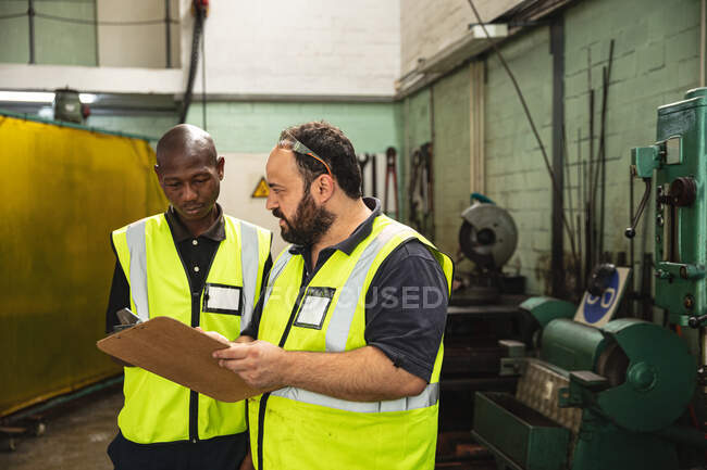 African American and Caucasian male factory workers wearing a high vis vest talking and holding clipboard. Workers in industry at a factory making hydraulic equipment. — Stock Photo