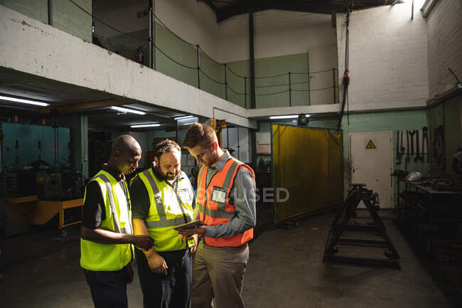 Deux ouvriers blancs et un ouvrier afro-américain portant une veste haute vis parlant et utilisant une tablette. Travailleurs de l'industrie dans une usine de fabrication d'équipements hydrauliques. — Photo de stock