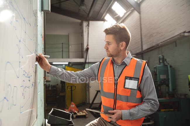 Caucasian male factory worker wearing a high vis vest holding tablet and writing on a board. — Stock Photo