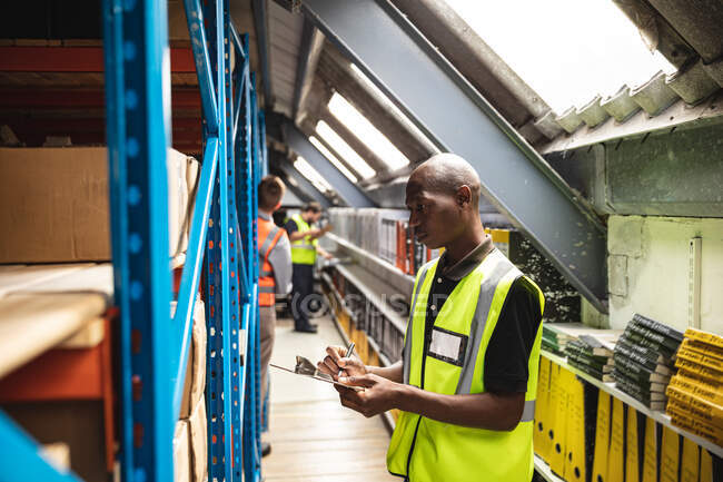 Two Caucasian and an African American male factory workers wearing a high vis vest talking and holding clipboard, checking stock. Workers in industry at a factory making hydraulic equipment. — Stock Photo