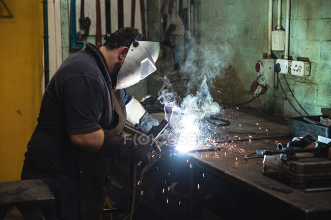 Trabajador de fábrica masculino caucásico que usa delantal oscuro y máscara de soldadura, de pie en un banco de trabajo, soldadura. - foto de stock