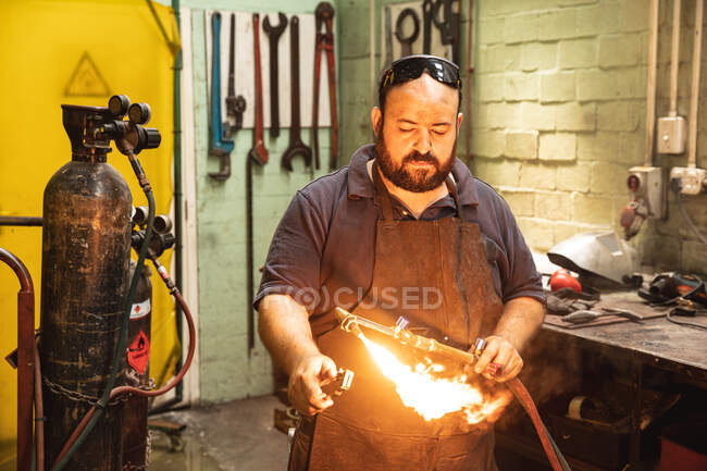 Trabajador de fábrica masculino caucásico que usa delantal oscuro y gafas de seguridad, de pie en un banco de trabajo, soldando una parte hidráulica. - foto de stock